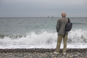 A gray-haired man with a backpack on his shoulder stands on the seashore