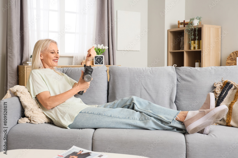 Sticker Mature woman massaging her wrist with percussive massager on sofa at home