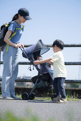 A mother pushing a stroller along a riverbank and her cute older brother taking care of and looking...