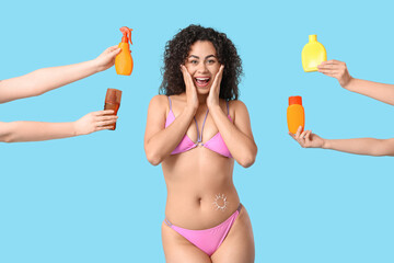 Surprised young African-American woman in swimsuit and hands with bottles of sunscreen cream on blue background