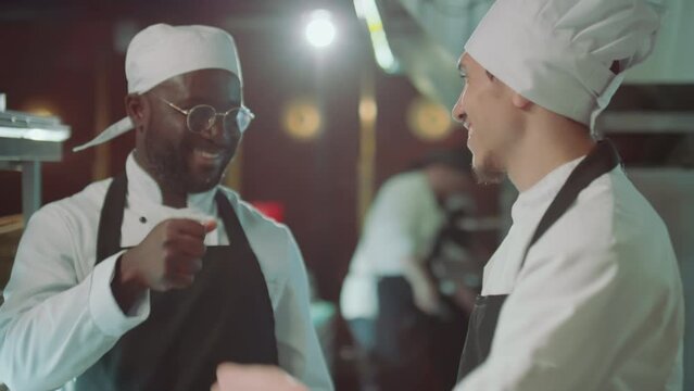 Young Caucasian chef and his Black coworker cooking together in restaurant kitchen, smiling and bumping their fists together