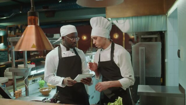 African American sous chef walking to colleague in restaurant kitchen, showing order on digital tablet and discussing job tasks
