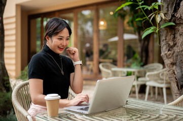 A cheerful woman sits at a table, looking at her laptop with a happy face, celebrating good news.