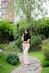 A positive, beautiful Asian woman talking on the phone while walking in a beautiful green garden.