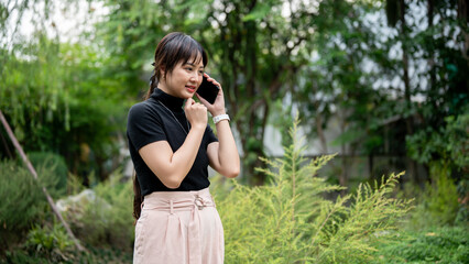 A positive, beautiful Asian woman talking on the phone while walking in a beautiful green garden.
