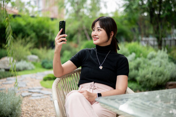 A beautiful Asian woman sits at a table in a backyard relaxing and using her phone.