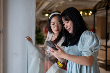 Two Asian women chat happily in a shopping mall corridor, looking at a smartphone together.