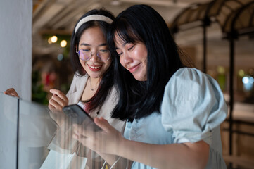 Two Asian women chat happily in a shopping mall corridor, looking at a smartphone together.