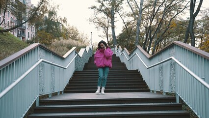 Smiling pretty woman in stylish jacket talking on the smartphone while walking down the stairs in the park. Seasonal style, autumn season concept. 