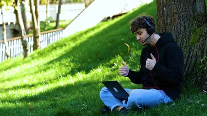 Happy man working with laptop and headset while making video calls, sitting on grass in the park. Autumn season. Education, working concept. Slow motion