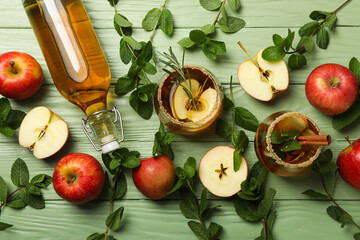 Glasses and bottle with apple cider, mint leaves and red apples on green wooden background, top view