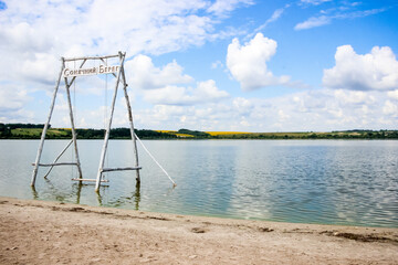 wooden swing in the water on a summer lake, beach, sand,  white clouds, reflection