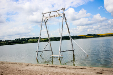 wooden swing in the water on a summer lake, beach, sand,  white clouds, reflection