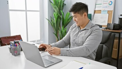 Smiling asian man using smartphone and laptop in modern office setting communicates productivity and connectivity.