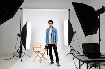 Portrait photo of young smart Asian professional photographer standing on white photo backdrop indoor studio set with set of strobe flash and soft box preparing for product and model shoot.