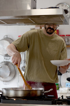 man slightly obscured, almost unrecognizable by the stainless steel fume hood, stirring a pan, green caci t-shirt, pots hanging in the background, kitchen environment.