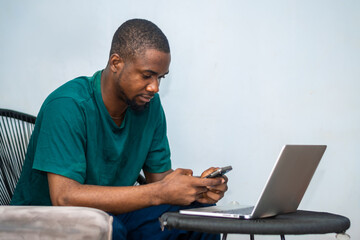 photo of young African American guy working on laptop and using smartphone. African man wearing green shirt, Texting, Internet, Technology. Mobile phone