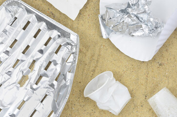 Silver tray, aluminium trash and white plastic and paper on beach after barbecue or party. Sand in...