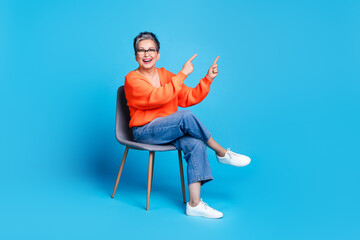 Full body photo of lovely senior lady sit chair point empty space dressed stylish orange garment isolated on blue color background