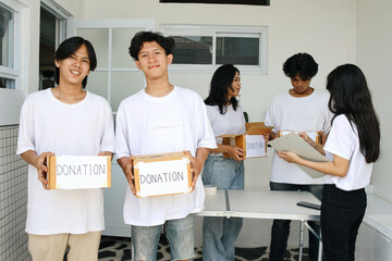 Group Of Donation Volunteer. Two Young Man Holding Donation Box Looking At Camera. Volunteers Helping People In The Backgound
