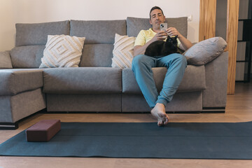 Man with Smartphone and Black Cat After Yoga Session in Living Room. Man with smartphone relaxes on...
