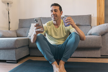 Man takes break from guided meditation on yoga block in living room, smiles while checking phone,...