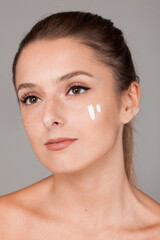 Young beautiful woman with healthy skin taking care of face with cream isolated on gray background...