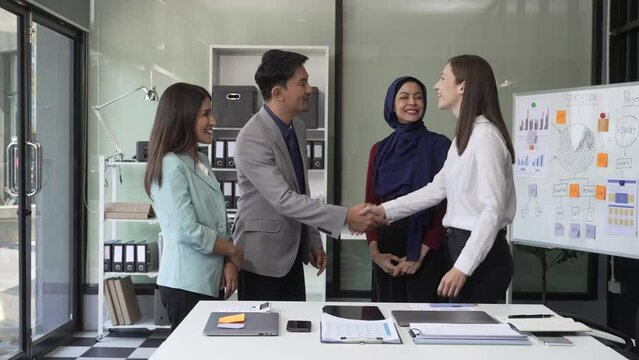A diverse team, including a middle-aged Asian businessman, Caucasian young businesswoman, Muslim hijab-wearing middle-aged businesswoman, meets in a boardroom to close an Annual General Meeting.