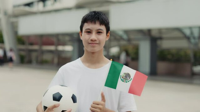 Portrait of a soccer fan holding the Mexican flag outdoors. A soccer player stands with a ball and the flag, gazing into the camera. Football and People Concept