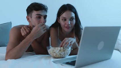 An affectionate couple watches a movie on a laptop and eats popcorn together in bed at night, sharing a cozy and intimate moment.