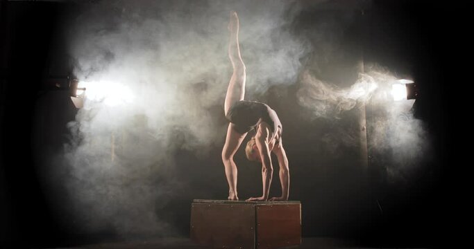 Acrobat in backbend on box raise leg up, stage performance in smoky spotlights