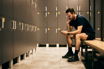 A sportsman sitting in a locker room, using a mobile phone, preparing for a workout.