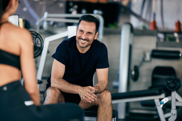 Focus on the smiling fitness man talking to a fitness girl, sitting on the bench, at the gym.