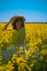 Portrait of beautiful woman in green dress, straw hat, with blooming yellow rapeseed field in background. Nature relaxation, style, and aesthetic.