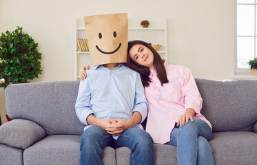 Love couple sits on sofa emotions man in paper bag with drawn smile woman smiling emotions of joy....