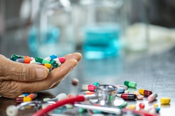 A close up of a hand holding a handful of pills with a stethoscope on the table.