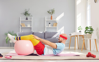 Fototapeta premium Funny young man in sportswear having home workout lying on floor on yoga mat with fit ball. Sporty male person exercising at home. Sport, fitness, health and home training concept.