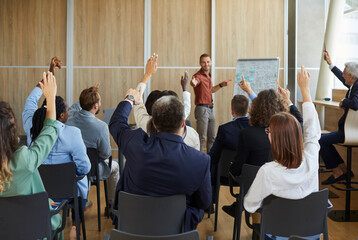Young colleagues raising hands to ask questions during business meeting in office. Diverse business people voting at the conference in meeting room listening their young male speaker.