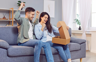 Delivery and online shopping. Couple excitedly and curiously opens box and rejoices in pleasant surprise of receiving new package. Couple is sitting on sofa and looking at contents of cardboard box. 