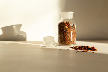 Orange and brown coloured dried spices and leaves in small open glass bottle and scattered on table in ray of sunlight
