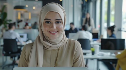 Focused Muslim female using laptop in contemporary office setting. Account specialist smiling while having productive day. Colleagues working on computers in background.