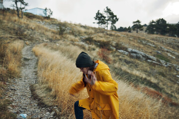 Woman in a vibrant yellow raincoat skateboarding down a dusty mountain road surrounded by nature...