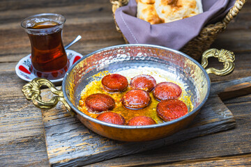 Turkish sausage fried in a copper pan, served with tea and bread, wooden background, close-up.
