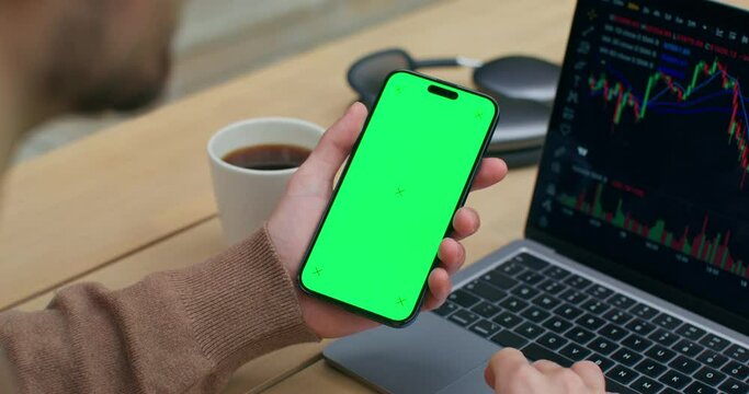 The man uses a mobile phone with green screen. He uses the laptop sitting at his desk. Graphs on the screen. An unrecognizable person