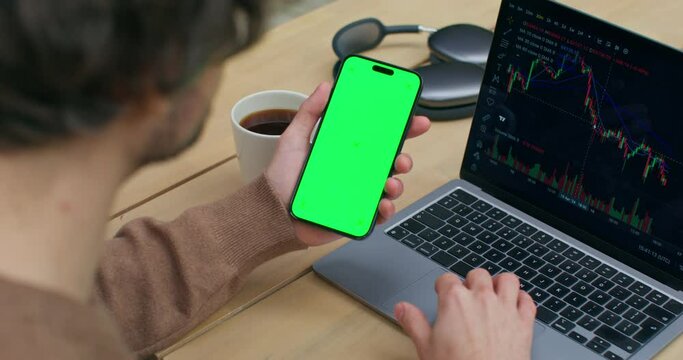 The man uses a mobile phone with green screen. He uses the laptop sitting at his desk. Graphs on the screen. An unrecognizable person