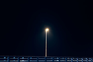 Solitary lamppost casts dim cold blue glow along shore pier standing against pitch black canvas of...