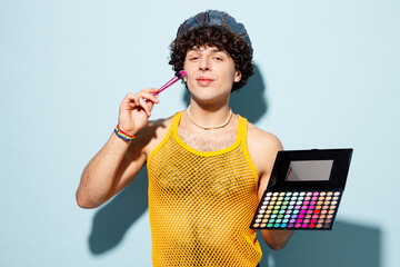 Young happy gay Latin man wears mesh tank top hat clothes do make up use brush hold palette of blushes isolated on plain pastel blue background studio portrait. Pride day June month love LGBT concept.