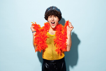 Shocked excited young happy gay Latin man wears mesh tank top hat yellow clothes tinsel posing isolated on plain pastel light blue background studio portrait. Pride day June month love LGBT concept.