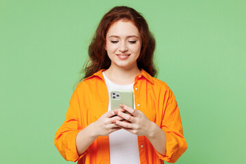 Young smiling ginger fun woman she wear orange shirt white t-shirt casual clothes hold in hand use mobile cell phone chatting online isolated on plain pastel light green background. Lifestyle concept.