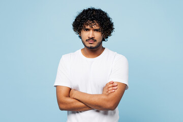 Young sad disappointed Indian man he wearing white t-shirt casual clothes hold hands crossed folded look camera isolated on plain pastel light blue cyan background studio portrait. Lifestyle concept.